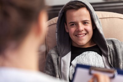 Boy with hood up smiles as he looks at a woman writing on a clipboard